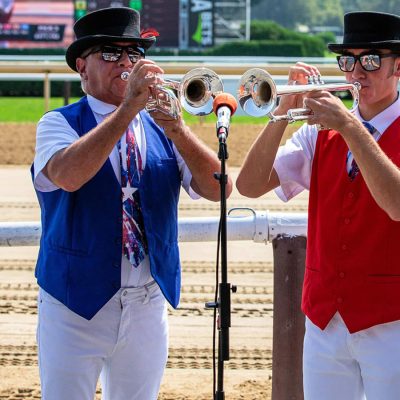 Two buglers in top hats and sunglasses, wearing blue and red vests over white shirts, play trumpets into a microphone at Saratoga Race Course, with the dirt track and rail visible behind them.
