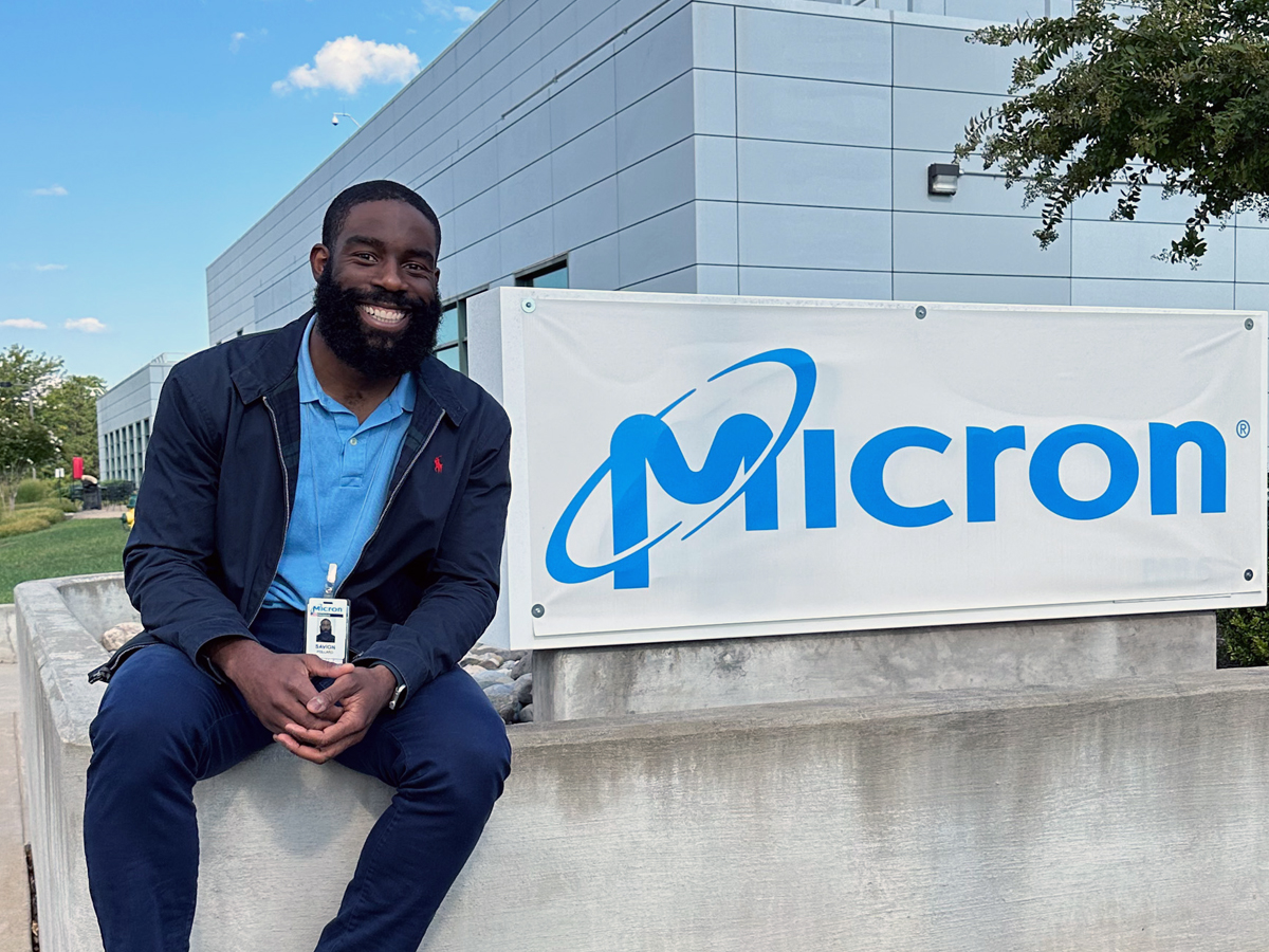Smiling man with Micron ID badge sits by a Micron sign outside company building.