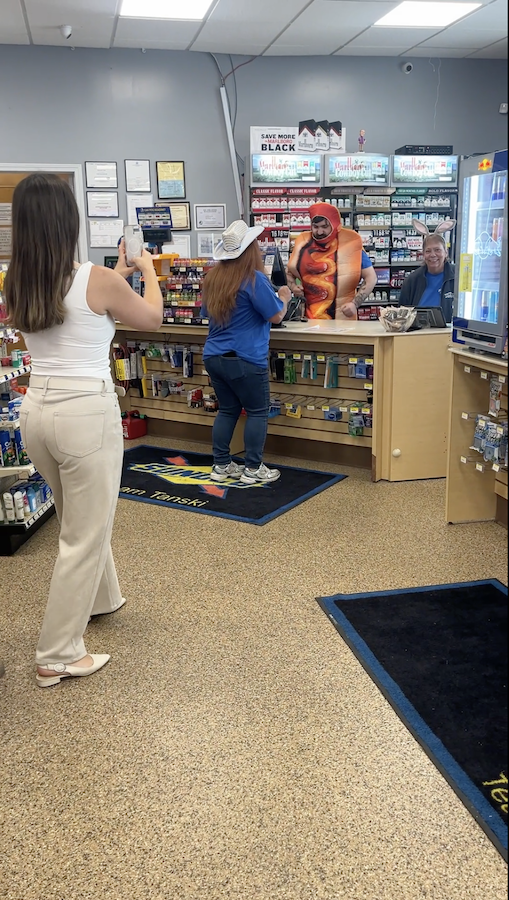 Person recording a customer and store employee in costume at a convenience store checkout counter.