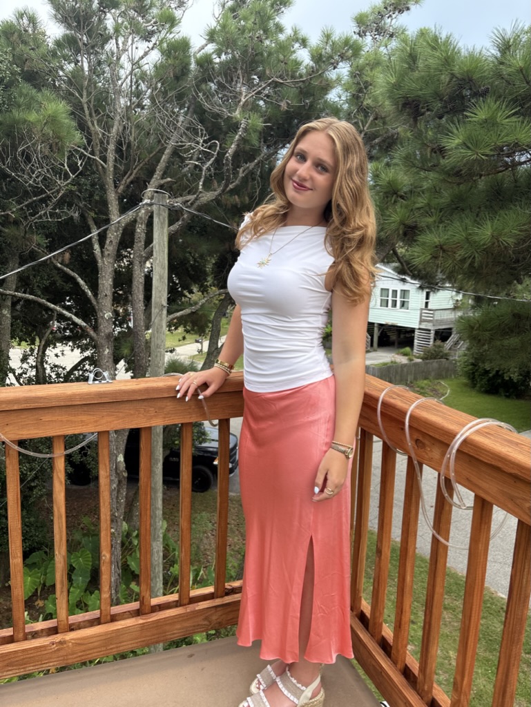 Person standing on a wooden deck outdoors, wearing a white top and pink skirt with trees in the background.