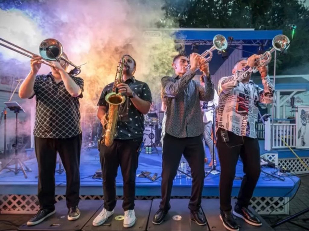 A four-piece horn section — trombone, saxophone, and two trumpets — performs on an outdoor stage lit with colorful smoke and haze under string lights.