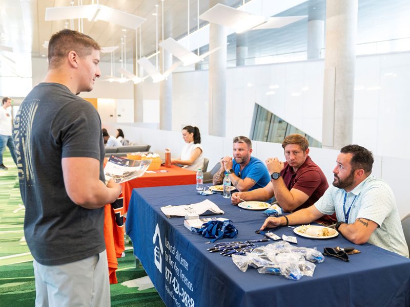 Three men seated at a National Call Center for Homeless Veterans table listen to a presenter in a flag-graphic T-shirt, with pens, lanyards, and pamphlets displayed on the navy tablecloth.
