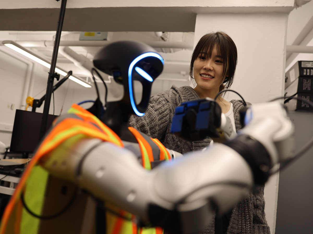 A person smiles while adjusting a humanoid robot in a safety vest.