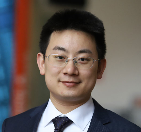 Headshot of a smiling man with glasses, wearing a dark suit, white shirt and patterned tie, with a blurred blue and orange background