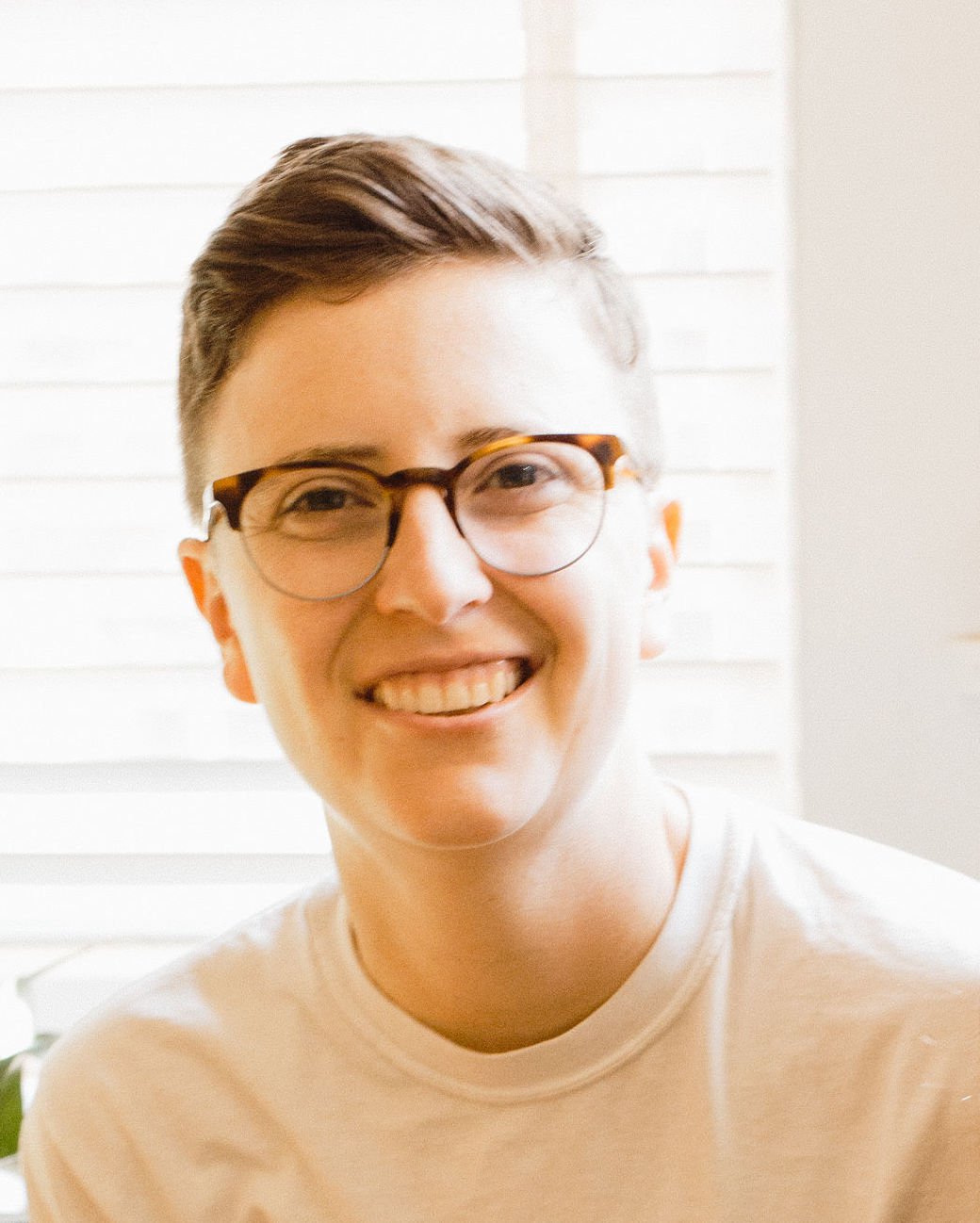 Person wearing round glasses and a light shirt, photographed indoors against a bright window background.