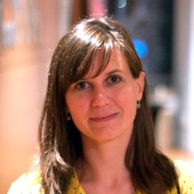 Headshot of a woman with long brown hair and bangs, wearing a yellow top, with a warmly lit indoor background