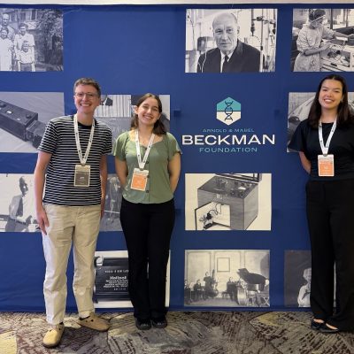 Three conference participants standing in front of a Beckman Foundation display featuring historical photos and scientific equipment.