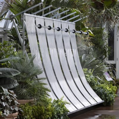 A curved metal water feature with vertical rivulets flowing down five arced steel bands sits inside a glass conservatory, surrounded by lush tropical foliage and ferns.