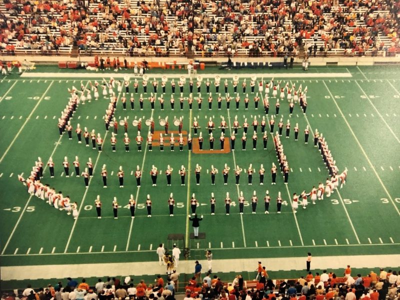 Marching band performs a large formation on a football field during a stadium halftime show, with spectators filling the stands.