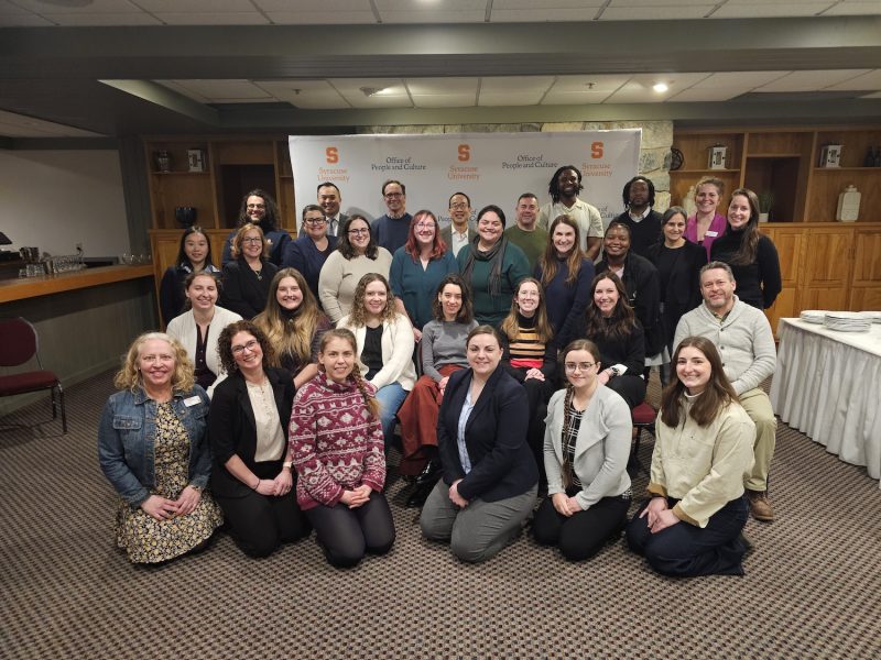 Group photo of approximately 30 Syracuse University Office of People and Culture staff members gathered in a meeting room, posed in several rows in front of a branded backdrop.
