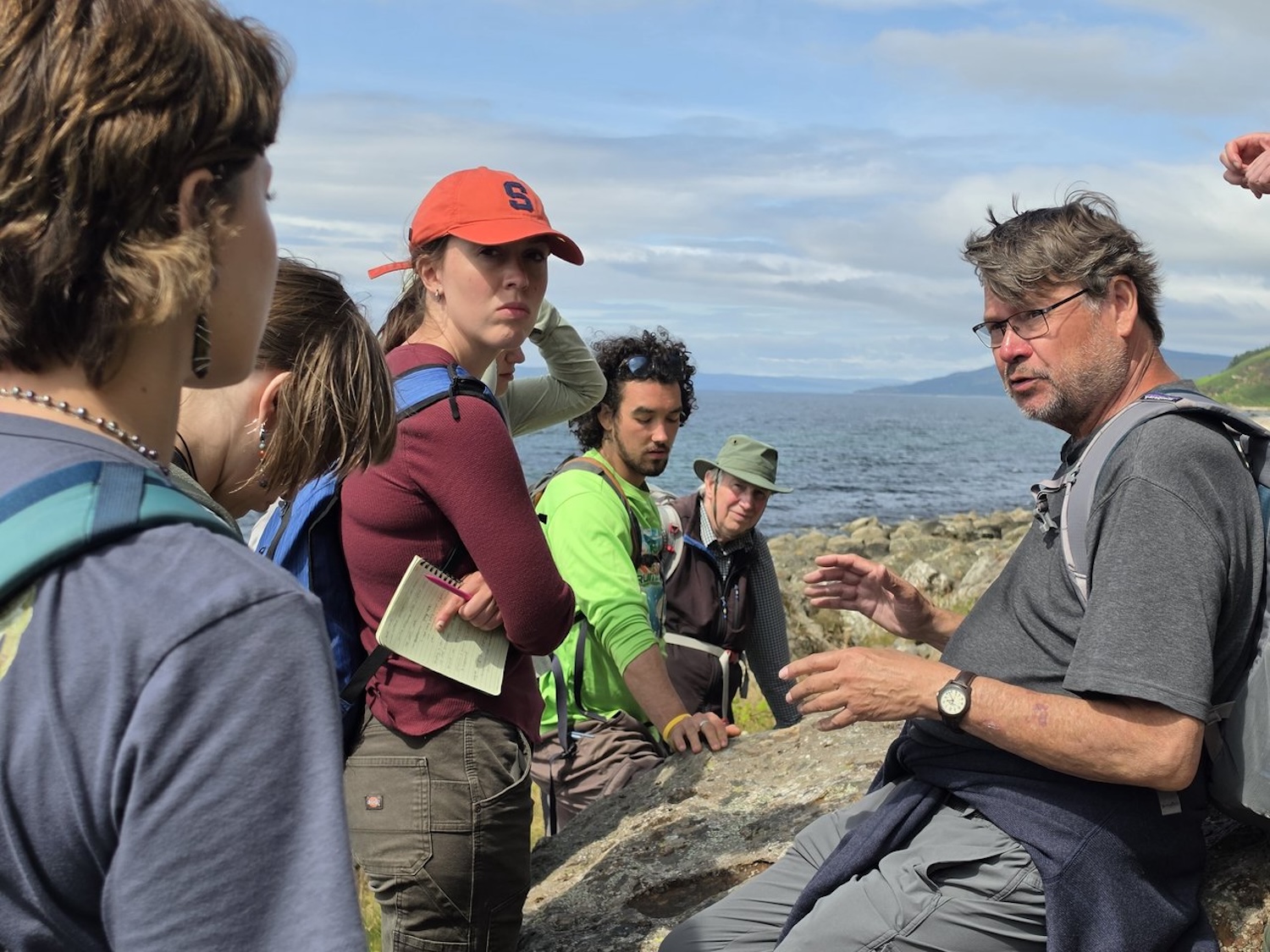 Field group listening to a guide on a rocky shoreline beside a large body of water.