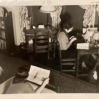 Historic black-and-white photo of two people in a bedroom-study, with one reading in the foreground and another typing at a desk beside bookshelves and lamps.