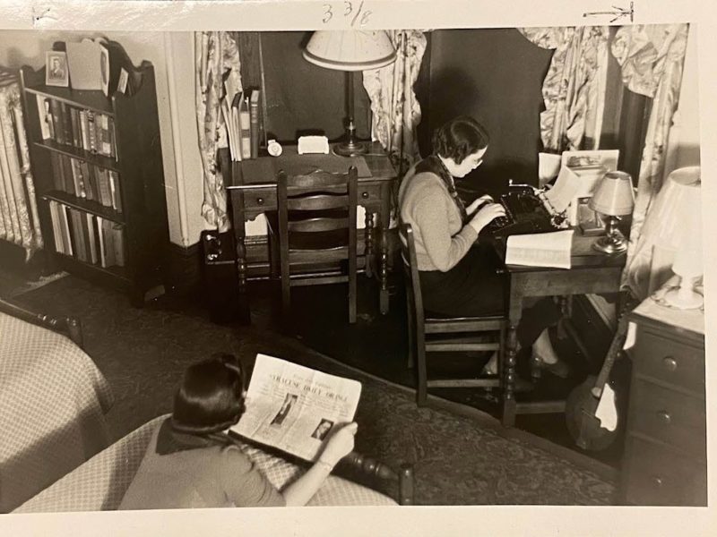 Historic black-and-white photo of two people in a bedroom-study, with one reading in the foreground and another typing at a desk beside bookshelves and lamps.