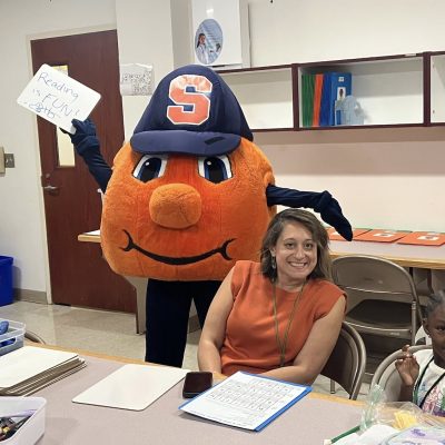 Syracuse mascot Otto the Orange holds a sign reading “Reading is FUN!” while visiting a classroom with participants seated at activity tables.