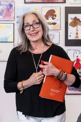Tere Paniagua smiles while holding an orange Syracuse University book, standing in front of a wall of children's artwork at La Casita.