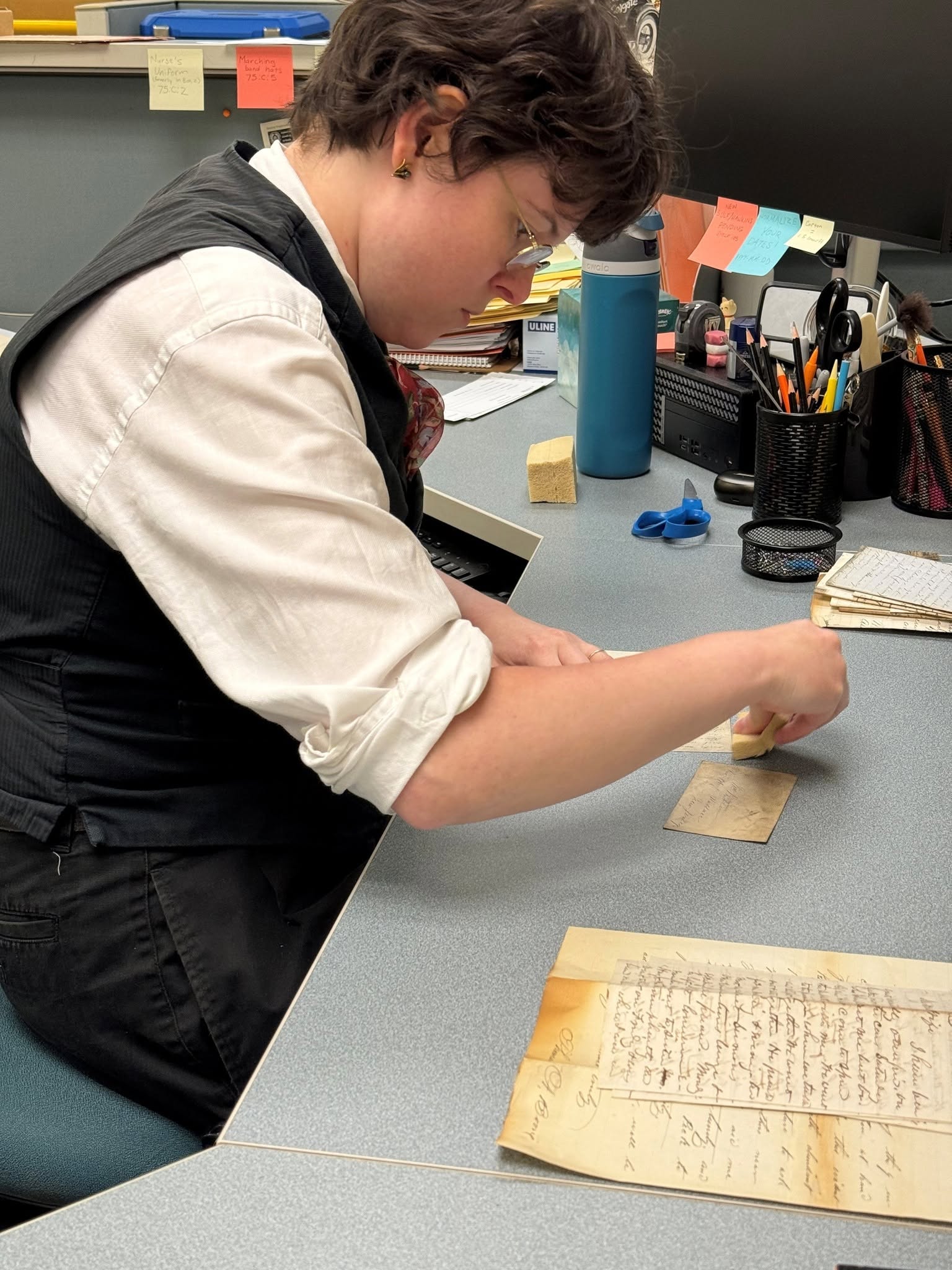 Person seated at a desk carefully cleaning handwritten historical documents with a sponge in an archival workspace.