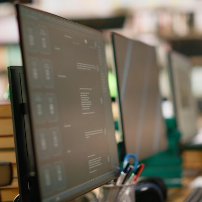 Row of computer monitors on a desk beside stacked books and a green reading lamp in a library setting.