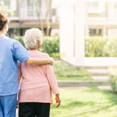 Caregiver walks beside and supports an older adult outdoors.