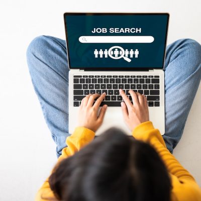 Person sitting cross-legged using a laptop displaying a job search screen with a search bar and candidate selection icon.