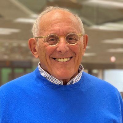 Person wearing round glasses and a bright blue sweater, standing in an office with a blurred background.