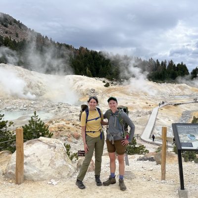 Two people standing near steaming geothermal features and boardwalks in a volcanic landscape.