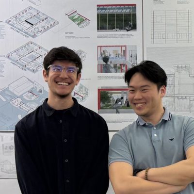 Two people standing in front of architecture presentation boards displaying building renderings, diagrams, and design plans.