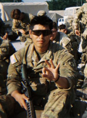 A soldier in desert camouflage and sunglasses gestures toward the camera while seated with other soldiers, tents and vehicles visible in the background.