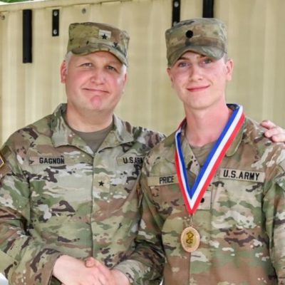 A U.S. Army general with his arm around a soldier wearing a medal, both in camouflage uniforms, smiling outdoors.