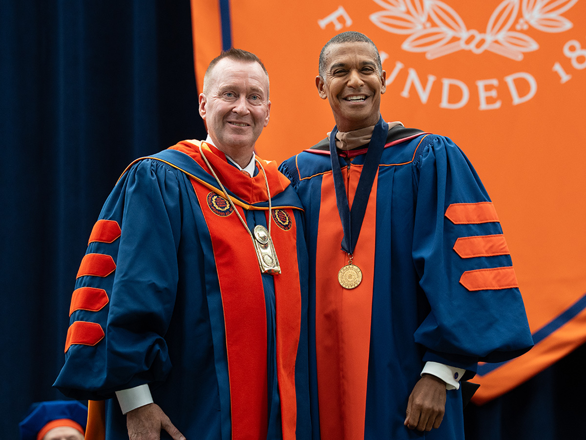 Two men in blue and orange academic regalia smile for a photo on stage, one now wearing the medallion around his neck, in front of an orange "Founded" banner.