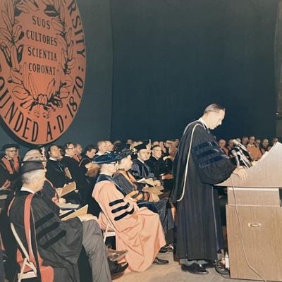 Academic ceremony with robed faculty seated onstage and a speaker at a podium beneath a large Syracuse University seal.