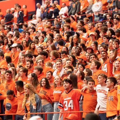 Syracuse fans in orange fill packed stands, cheering during a game.
