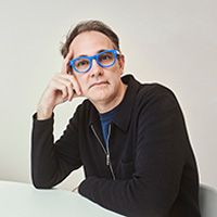 Headshot of a man with gray hair and blue-framed glasses, resting his chin on his hand, wearing a black jacket against a neutral background.