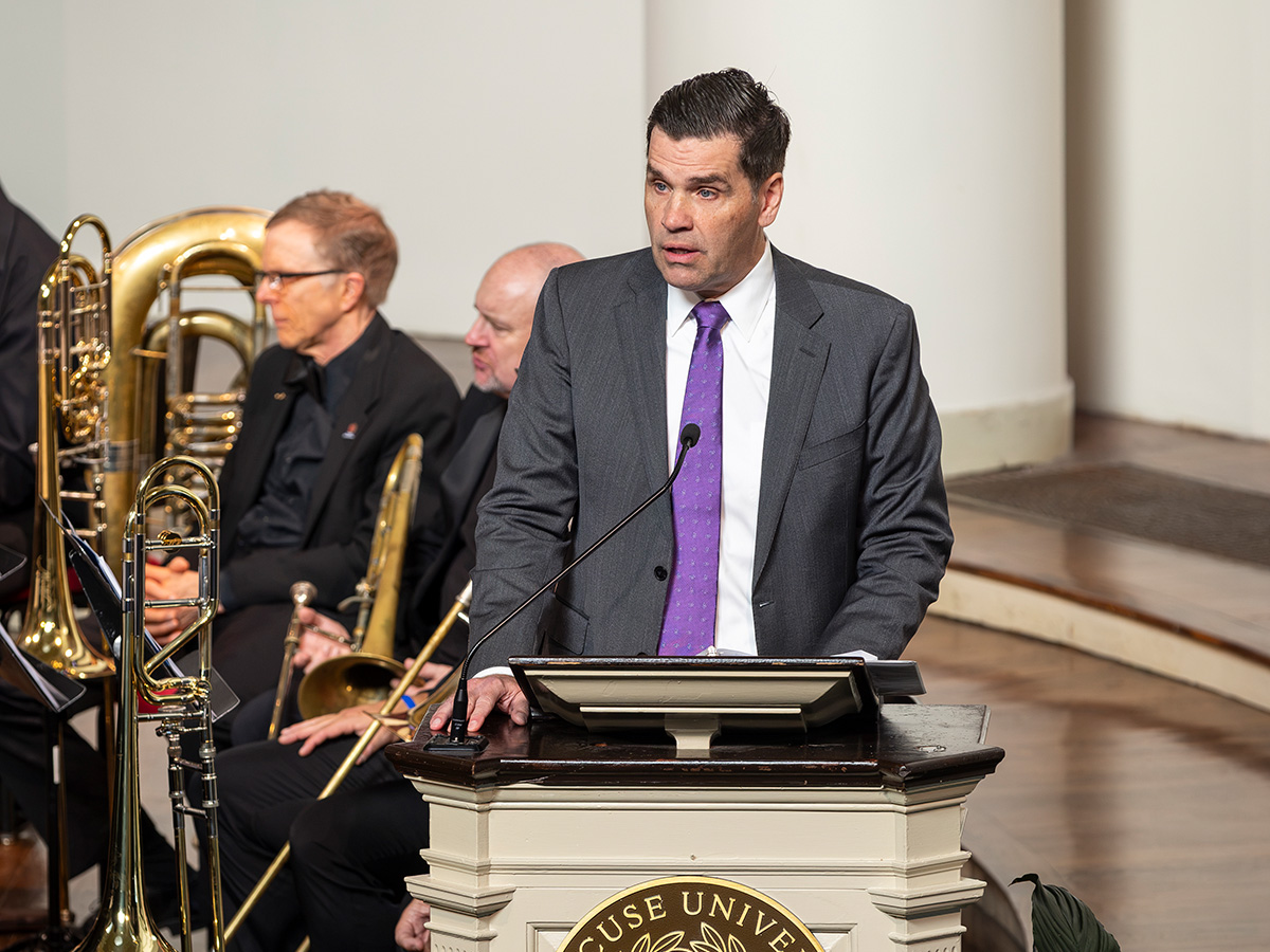 A man in a gray suit and purple tie speaks at a Syracuse University podium, with orchestra members and brass instruments visible behind him.