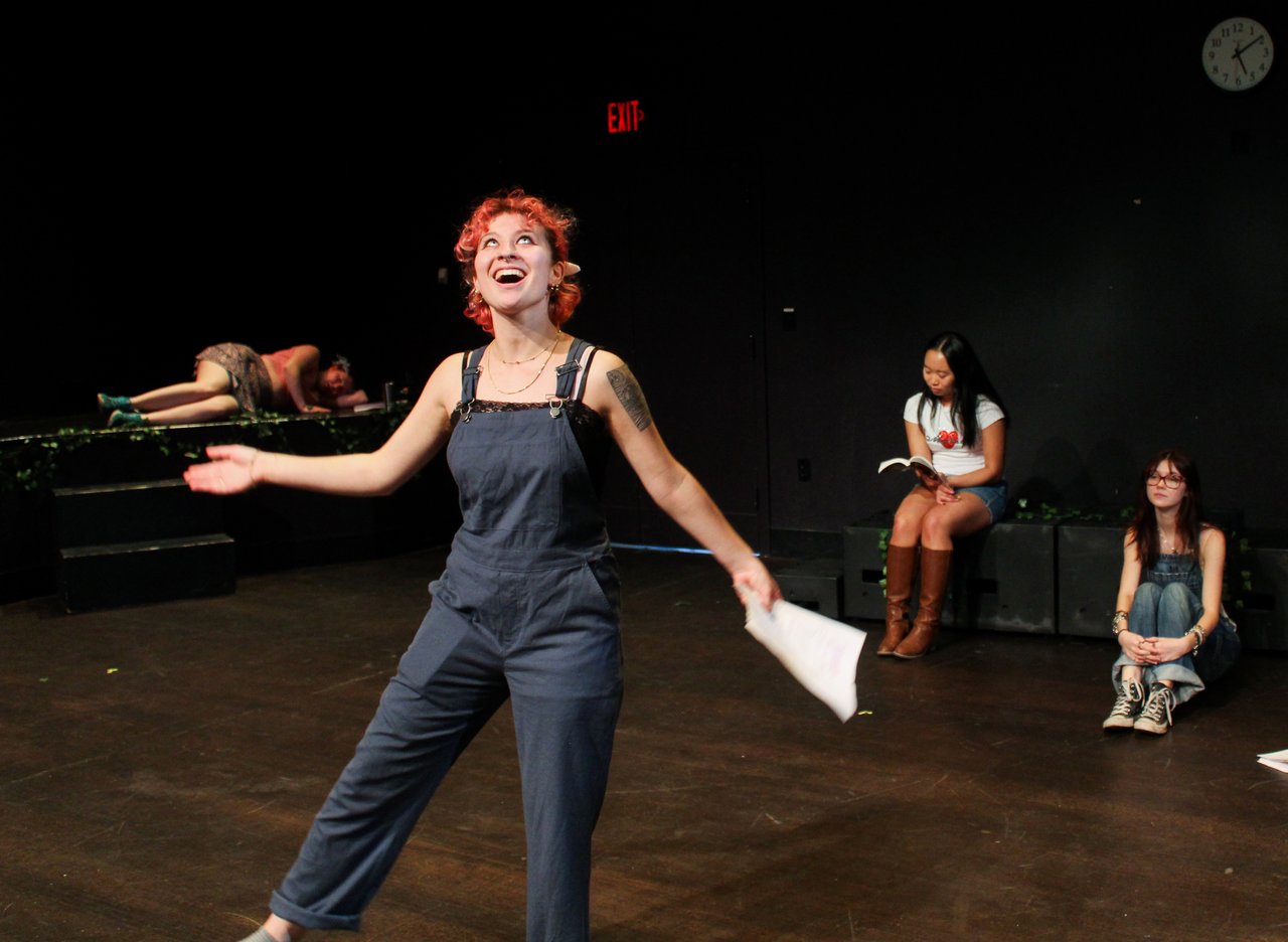 Student actor performs on a theater stage while others sit nearby during a rehearsal or workshop.