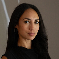 A woman with long dark hair wearing a black top, looking directly at the camera against a neutral background.