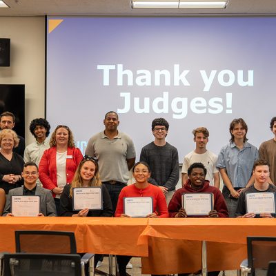 Winners of LaunchPad's 2026 Ideas Fest hold their first- and second-place certificates at a table in the front row, with judges standing behind them beneath a screen reading "Thank you Judges!"