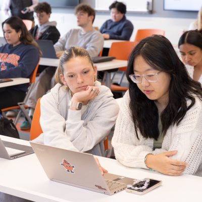 Students seated at tables in a classroom, working on laptops while following a lesson.