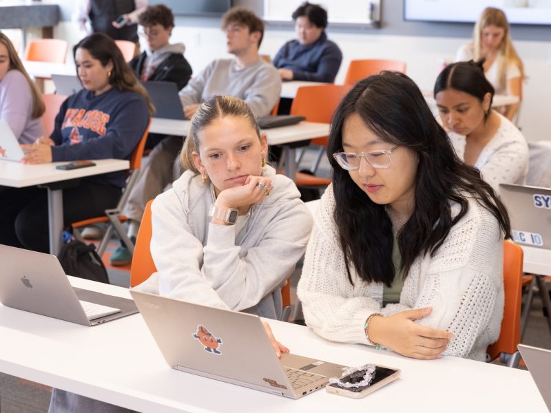 Students seated at tables in a classroom, working on laptops while following a lesson.