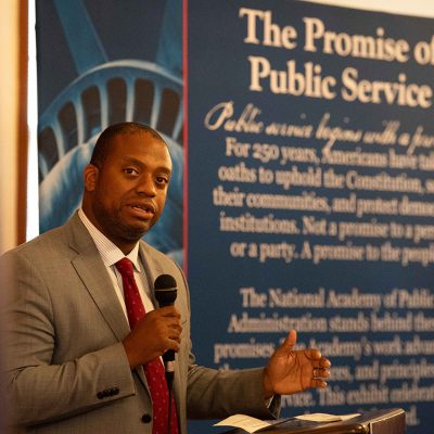 A speaker in a gray suit addresses an audience in front of a banner reading "The Promise of Public Service.