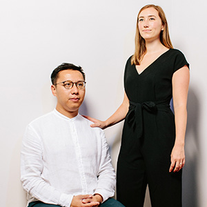 A seated man in a white shirt and a standing woman in a black jumpsuit, her hand on his shoulder, both facing the camera against a gray background.