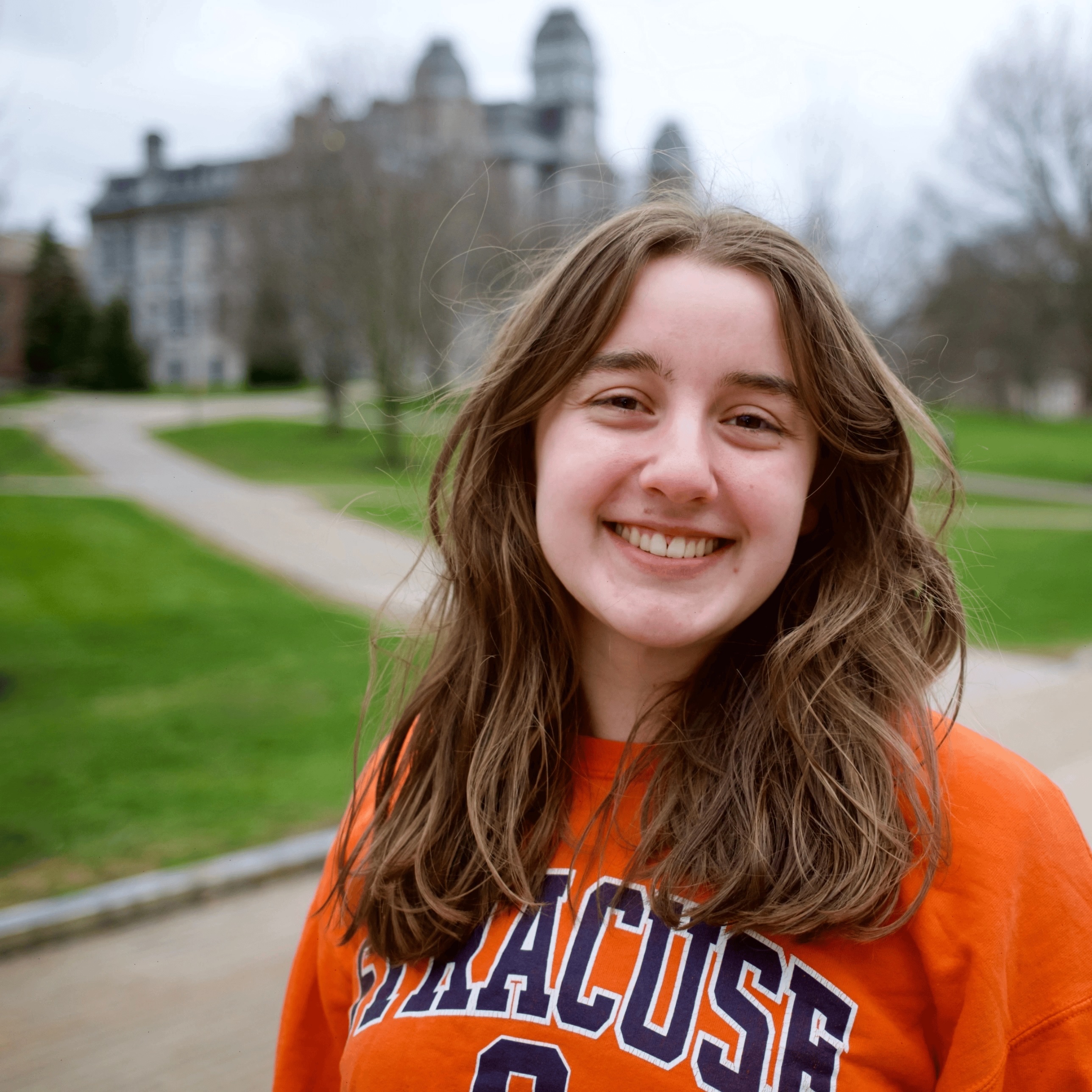 Person in an orange Syracuse shirt standing on a campus walkway with a blurred academic building in the background.
