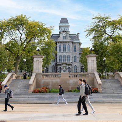 Students walk across the Syracuse University campus plaza in front of a historic stone academic building framed by trees and broad staircases.