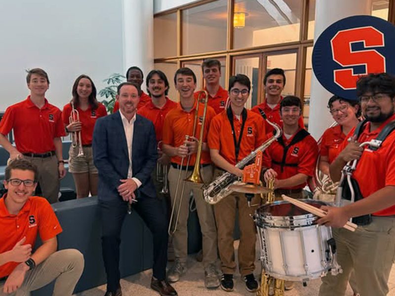 Syracuse University Marching Band members in orange polos pose with their instruments alongside a man in a navy suit, in front of a large Block S logo.
