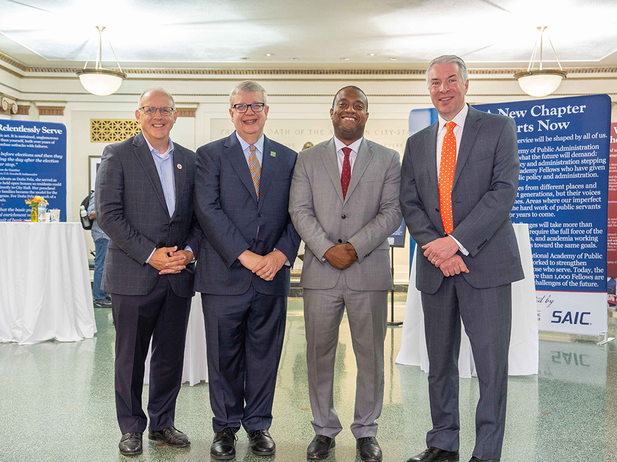 Four men in suits stand side by side and smile in a lobby, with blue exhibit banners behind them.
