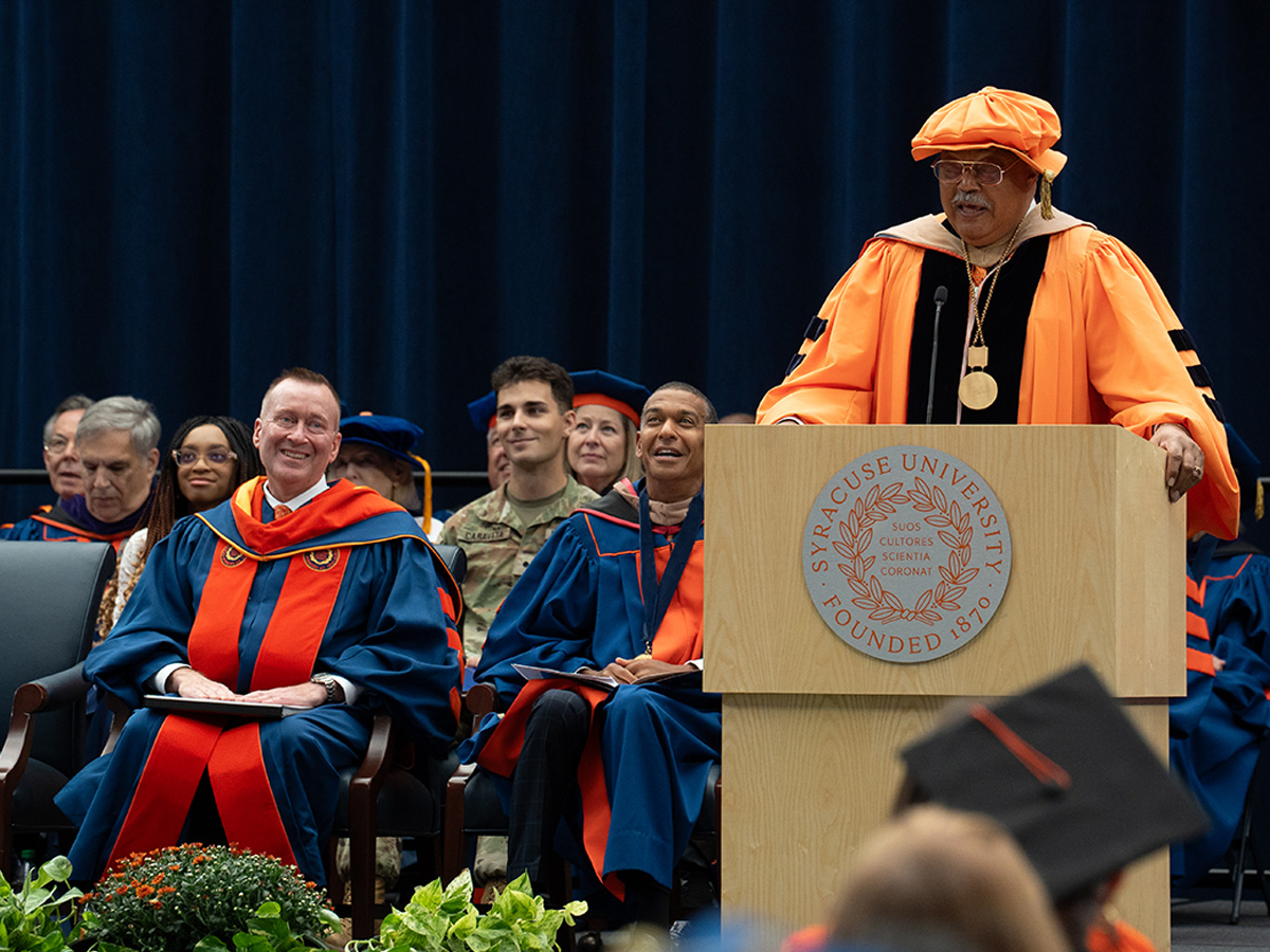 A speaker in orange academic regalia and a matching cap addresses the audience from a Syracuse University podium, while seated faculty and audience members in academic dress smile and look on.