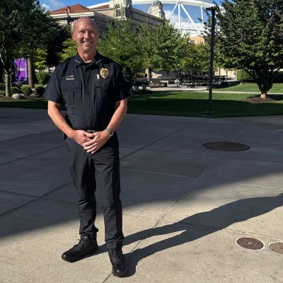 Michael Bunker, Chief of the Syracuse University Department of Public Safety, stands in his uniform on a campus plaza, smiling with hands clasped in front of him.