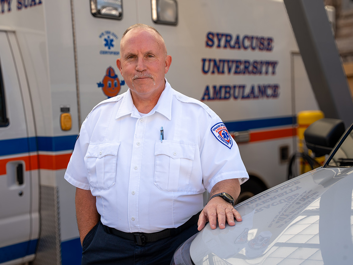 A man in a Syracuse University Ambulance uniform stands beside a vehicle, with a Syracuse University Ambulance vehicle visible in the background