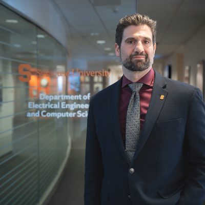 A man with dark hair, a beard, and a maroon shirt with a gray patterned tie under a navy blazer stands in a hallway, in front of a glass wall with text reading 'Syracuse University Department of Electrical Engineering and Computer Science.