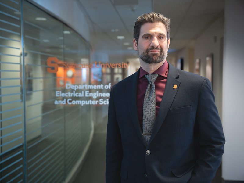 A man with dark hair, a beard, and a maroon shirt with a gray patterned tie under a navy blazer stands in a hallway, in front of a glass wall with text reading 'Syracuse University Department of Electrical Engineering and Computer Science.