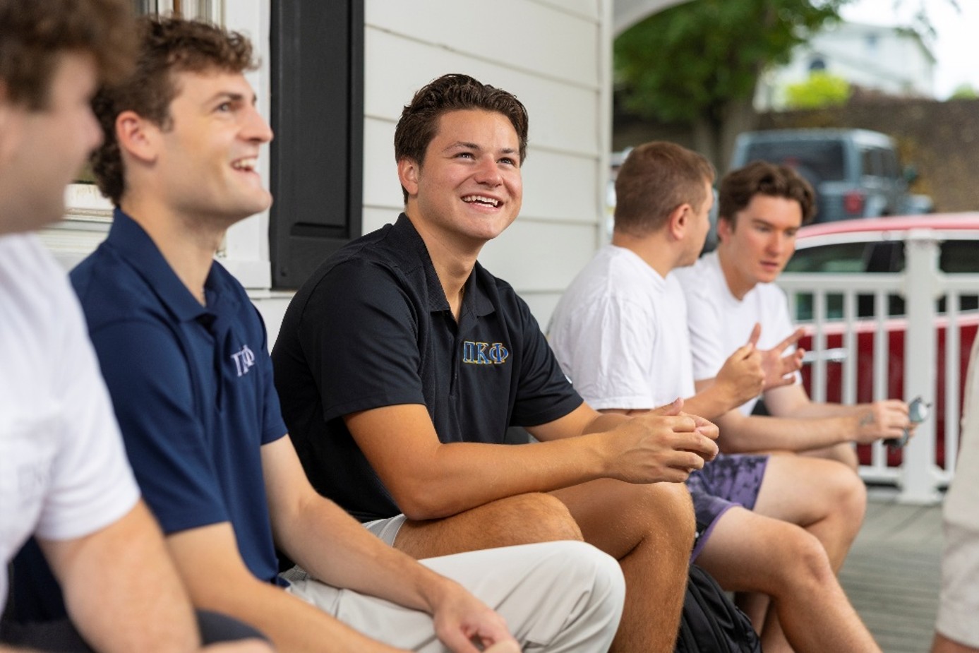 A group of people sit and laugh together on a porch, several wearing Pi Kappa Phi polo shirts.